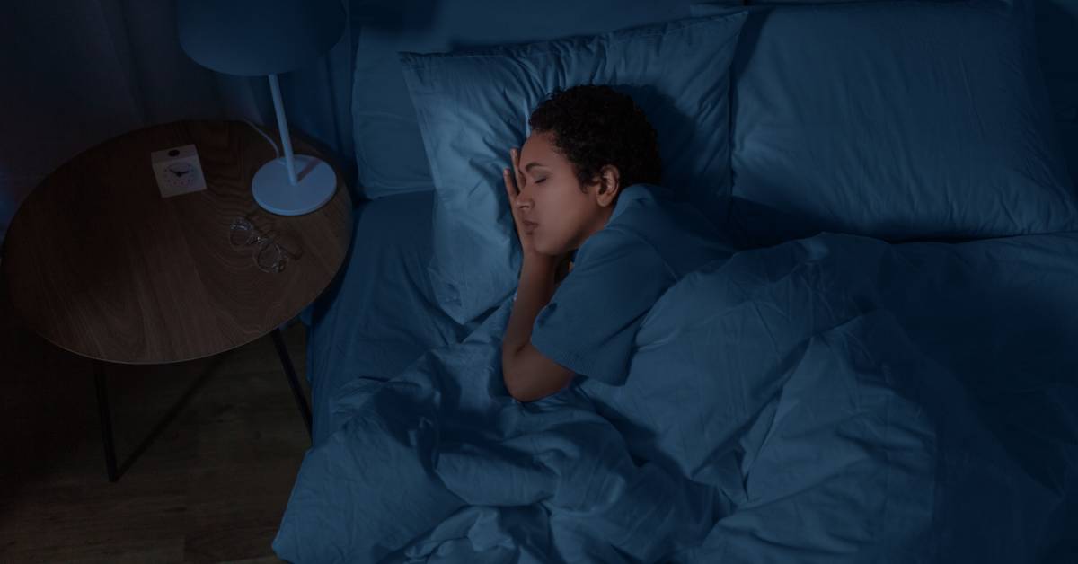 A person sleeping on their side with a relaxed expression in a dark bedroom. They have plump pillows and a puffy comforter.