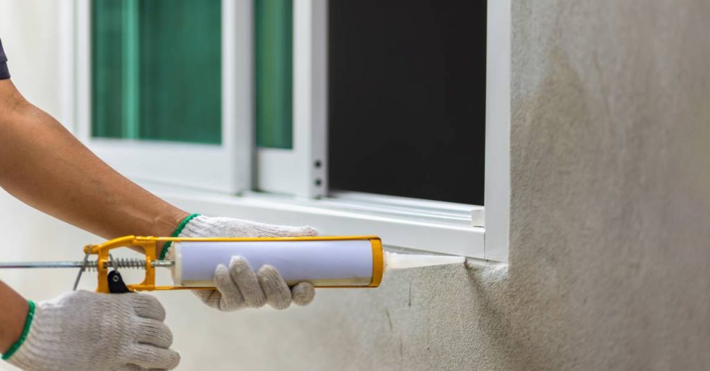 A person wearing gloves while using a caulking gun. They apply sealant to an exterior window frame on a beige stucco house.