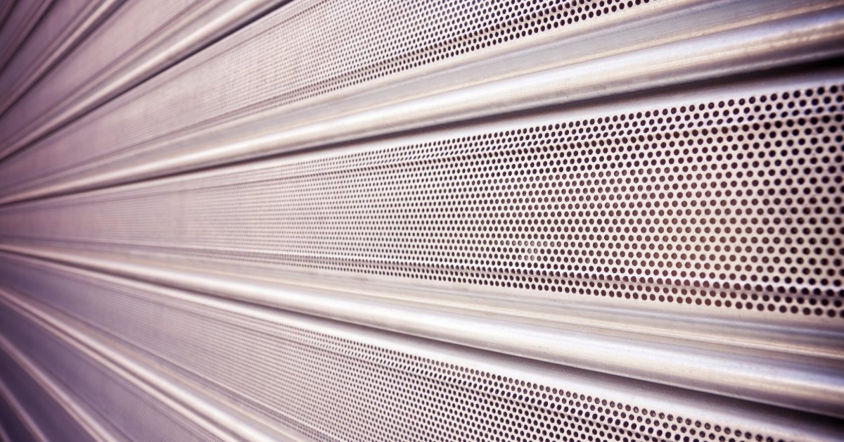 A close-up of the perforated slats of a silver security shutter. Nothing on the other side of the shutter is visible.