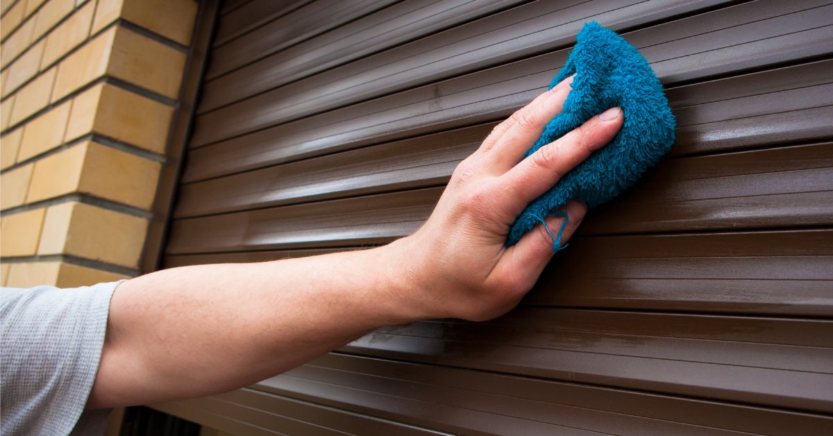 A brown rolling security shutter almost completely closed over a window. A person is wiping the shutter slats with a clean rag.