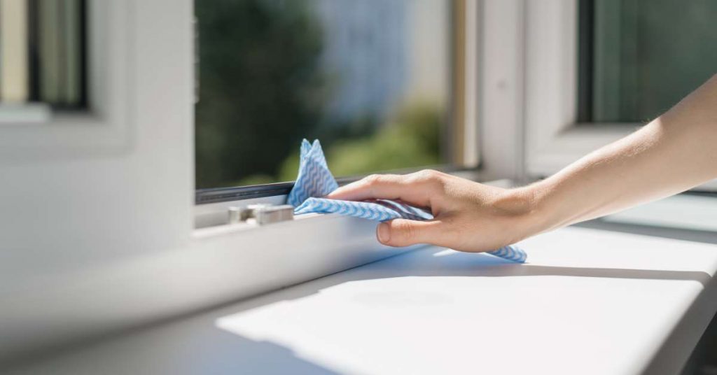 A person wiping the bottom of an open window's frame with a cleaning cloth. Sunlight illuminates the white windowsill.