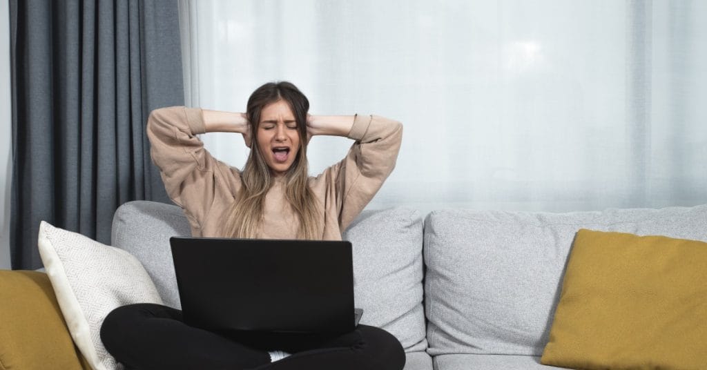 A woman sitting on a couch with her hands on her ears to block out noise. Behind her is a large window covered with curtains.