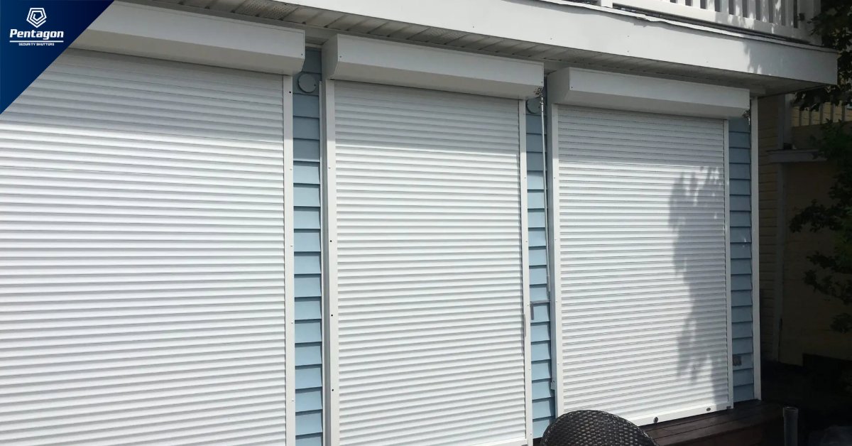 A row of white roll-down security shutters closed over three large windows on the exterior of a residential home.